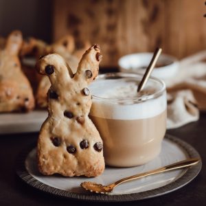 Häschen aus einem Joghurt Teig mit Schokotröpfchen und Karamelltröpfchen. Das Häschen leht an einem Glas mit einem Cappuccino. Häschen und Glas stehen auf einem Teller. Davor liegt ein Löffel. In dem Glas befindet sich ein goldener Strohhalm.